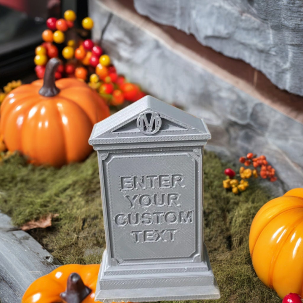 Personalized gothic tombstone displayed on moss-covered surface with decorative seasonal elements, featuring monogrammed crest and customizable text panel, shown in stone-gray finish with detailed architectural elements and ornate pedestal base