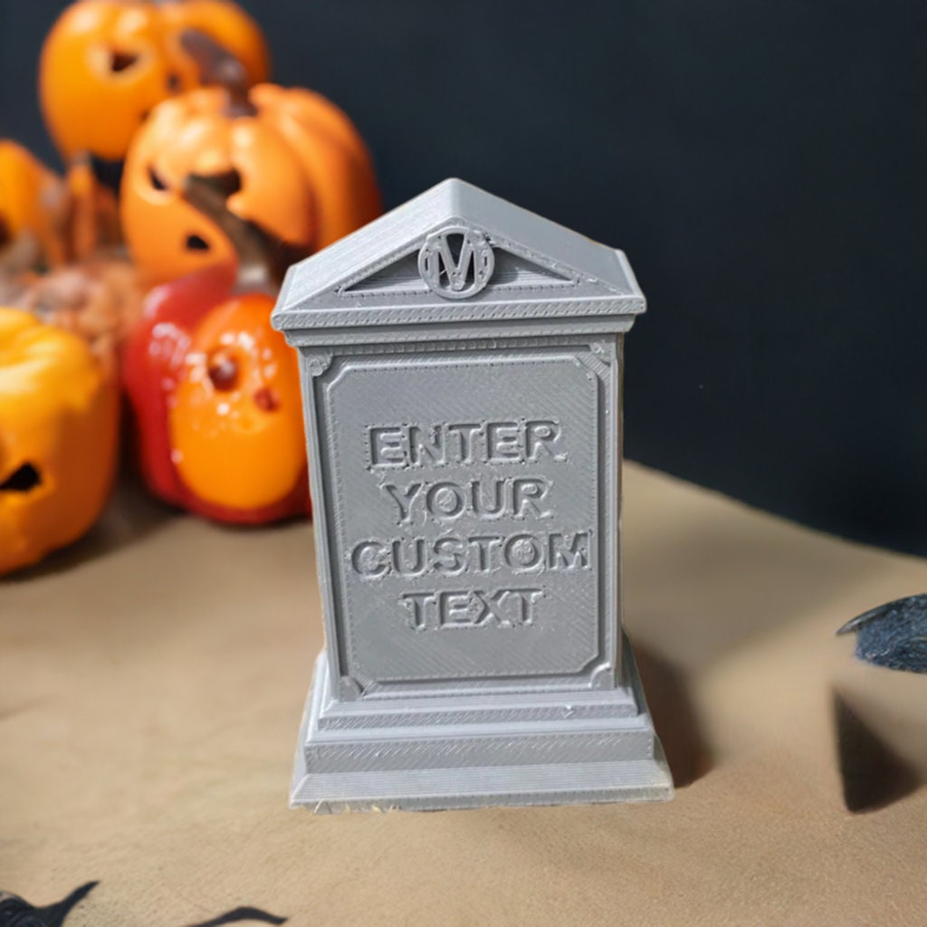 Gray 3D printed medieval tombstone with customizable text area and ornate crest, shown in miniature size with decorative border details and pedestal base, photographed with orange pumpkins in soft focus background