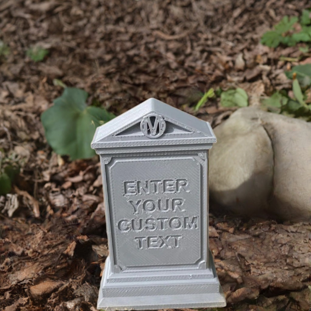 Custom gothic tombstone prop displayed in natural outdoor setting with mulch and green foliage, featuring decorative triangular crest and customizable text panel, shown in gray stone-like finish against natural rocks and leaves