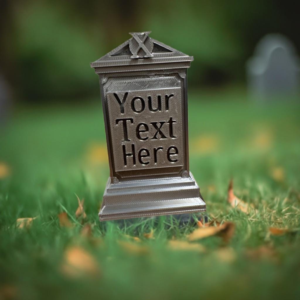 Miniature gothic tombstone displayed in lush green grass with autumn leaves, featuring customizable text panel and decorative monogram crest, shown in metallic gray finish with detailed architectural elements and bokeh effect background
